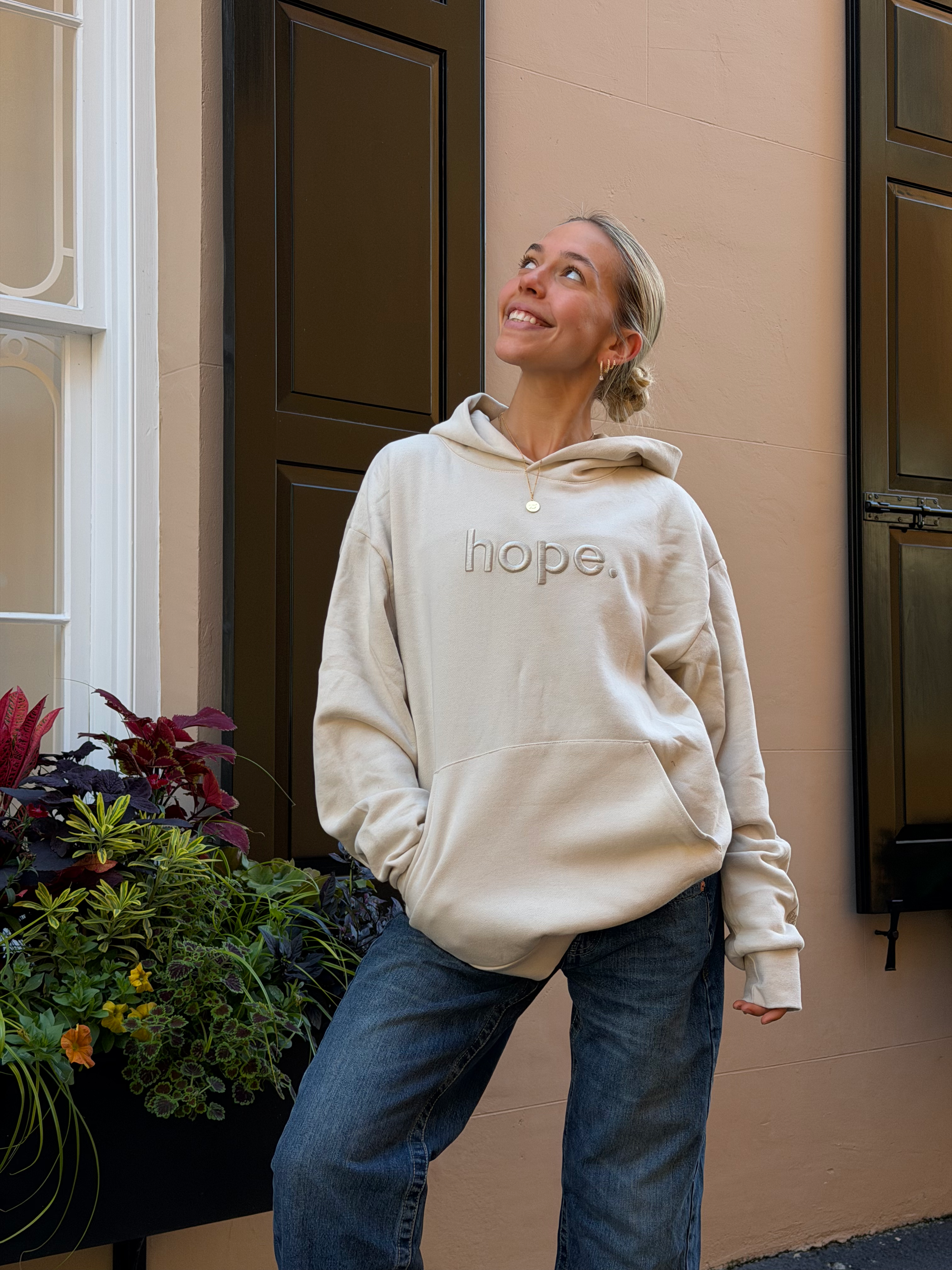 Person wearing a beige hoodie with 'hope' text, standing outdoors next to a building.